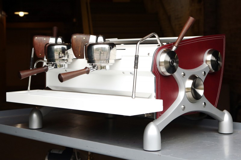 This image is a front-side view of the Slayer Espresso custom machine, leather sides, back/front panel and drip/cup trays powder coated white, x-legs powder coated silver, peruvian walnut wood accents, 2 groups at traditional height, manual dosing.